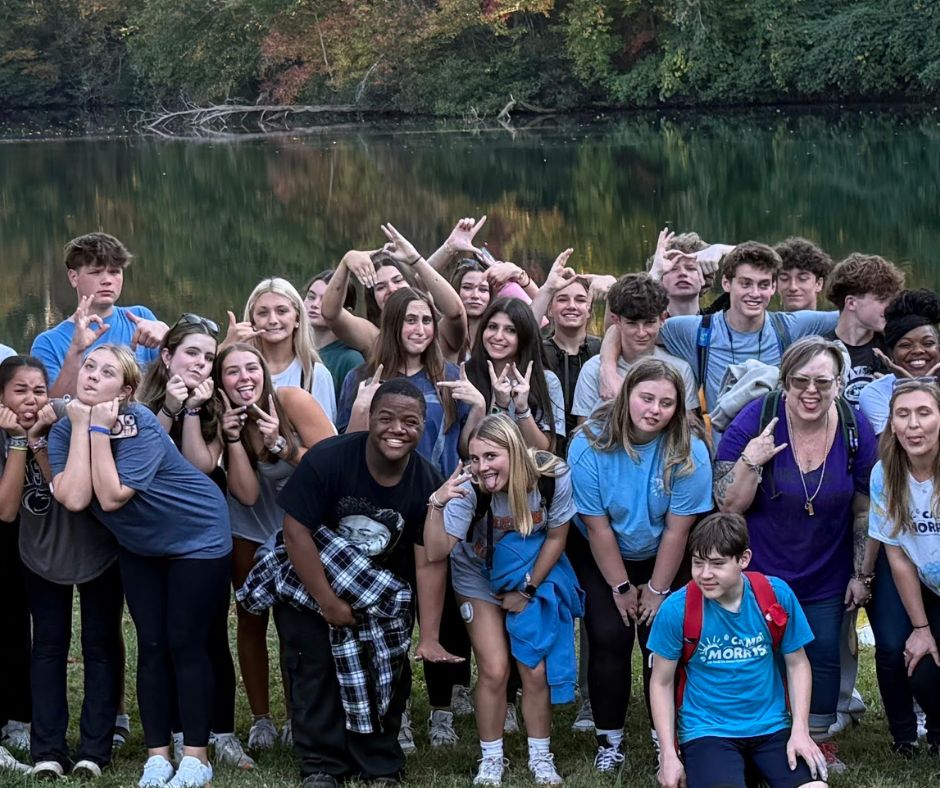 Group of teens and adults posing playfully by a lake, showcasing camaraderie and joy, reflecting the supportive community theme of The Diabetes Family Connection's Real Talk program for teens affected by T1D.